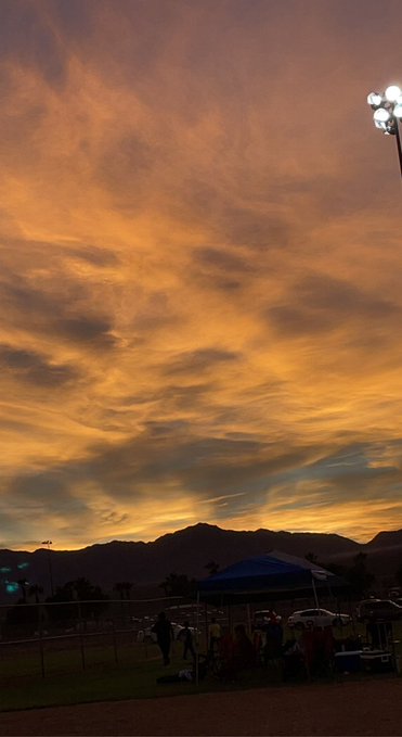 A dramatic sunset sky with orange and gold colored clouds over silhouetted mountains, featuring a stadium light pole and what appears to be a sports field with a blue tent below.