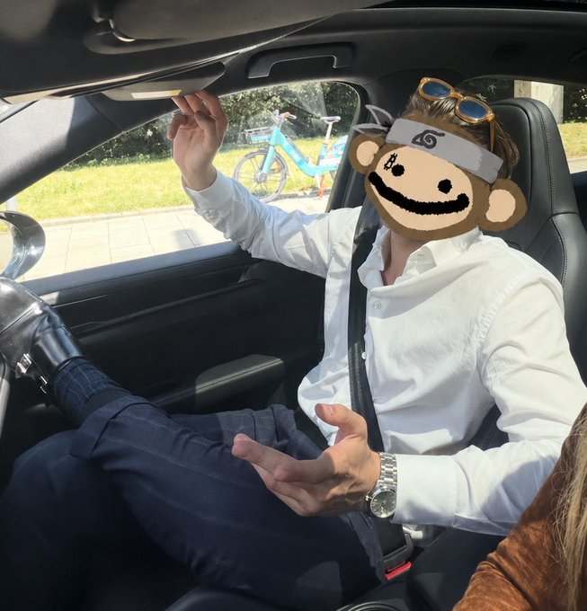 A man in a white shirt and tie sits in the driver's seat of a car, his face obscured by a monkey meme filter, gesturing with his hands while wearing sunglasses on his head and a silver watch.