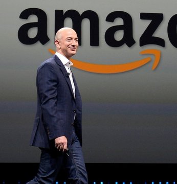 Jeff Bezos walks in a suit on a stage in front of a large screen displaying the Amazon logo.