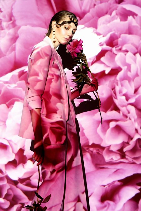 A fashion editorial photo of a woman wearing a pink satin suit, holding a pink peony flower against a backdrop of large pink flowers.