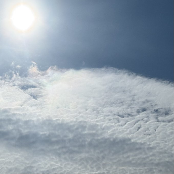 A bright sun shines over a thick layer of white clouds against a blue sky, with snow-capped mountains faintly visible in the distance.