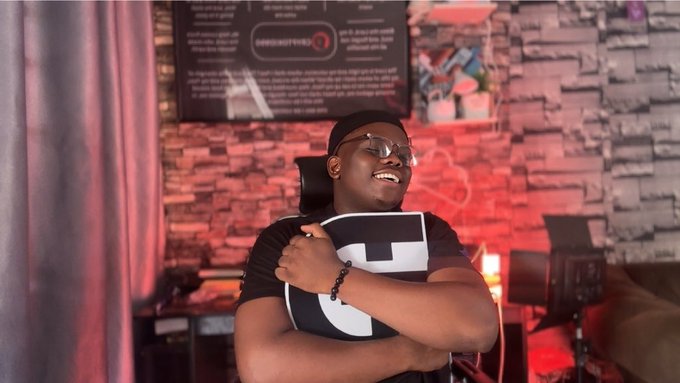 A person wearing glasses and a black and white t-shirt sits in a room with a brick wall and red lighting, laughing with their hand on their chest.