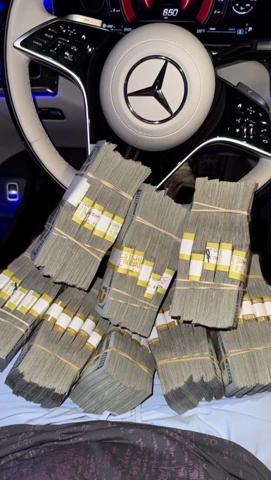 Interior shot of a Mercedes-Benz showing the steering wheel and dashboard, with several large stacks of US currency banded together in yellow wrappers piled on the seat.