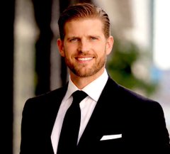 A portrait photograph of a smiling man with short blonde hair, wearing a dark suit, white shirt, and black tie, with a blurred indoor background.