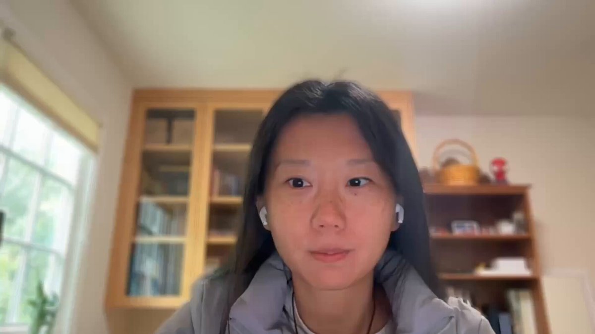 The image shows a woman wearing a grey jacket sitting at a desk, looking directly at the camera while holding an earbud in her right ear. The background features a window that allows natural light into the room. There are also books visible on the desk next to the woman.
