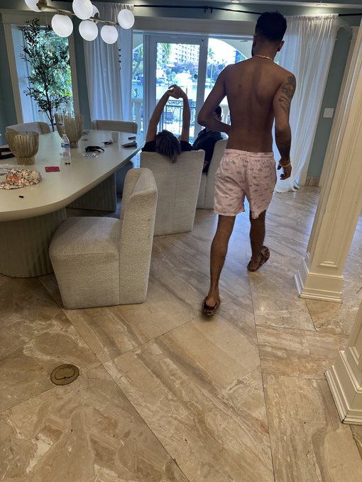 A shirtless man in patterned shorts walks through a modern living room while a woman reclines on a sofa in the background.
