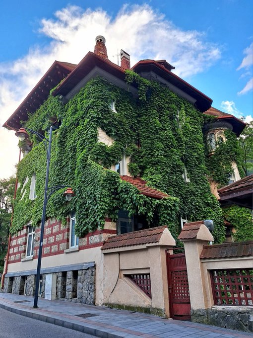 A picturesque European-style multi-story house entirely covered in lush green ivy with a red-tiled roof, decorative chimneys, and a stone-and-stucco perimeter wall with a wooden gate, captured under a blue sky with scattered clouds.