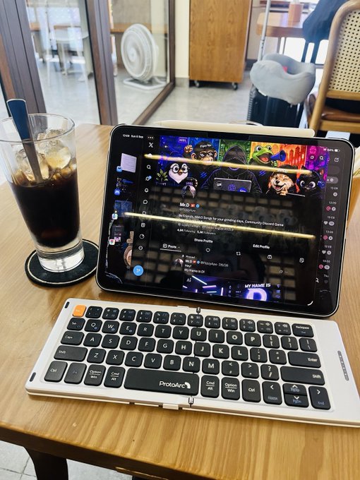 A wooden cafe table holds an iced coffee drink beside a tablet displaying a Discord community Discord server and a 'ProtoArc' foldable wireless keyboard.