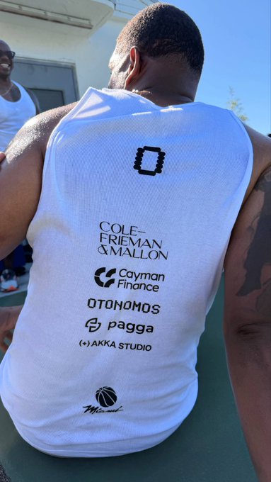 A man wears a white athletic jersey branded with sponsor logos including "Cole-Frieman & Mallon," "Cayman Finance," "Otonomos," "Pagga," and "Akka Studio," with the "Miami" logo at the bottom. The jersey relates to the post's mention of pickup games in Miami and NYC, highlighting the event's sponsorship branding while the player bends forward, likely during a game.
