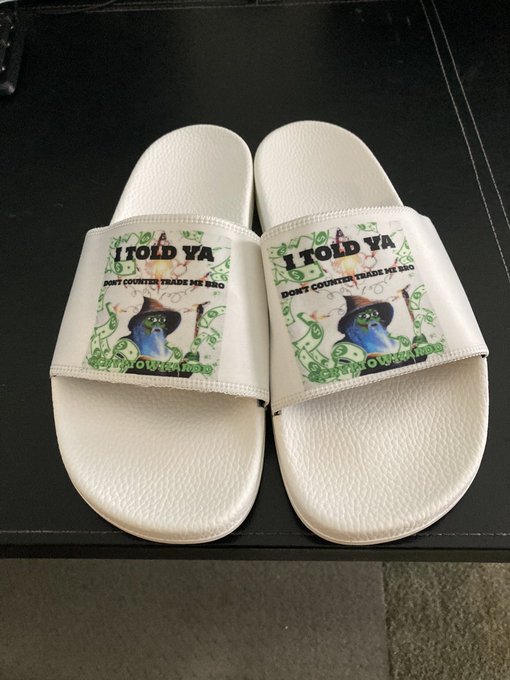 A pair of white slide sandals features a parody design mocking a $100 bill, with the text "I TOLD YA" and "DONT COUNTERFEIT TRADE ME BRO" surrounding a wizard figure in place of Benjamin Franklin—visually playing on the caption "Told you" by literally stating it on the design.