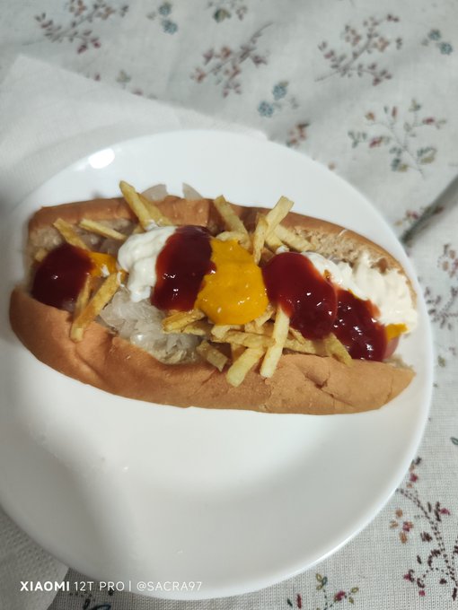 A hot dog in a bun topped with shoestring fries, mayonnaise, ketchup, and mustard, served on a white plate with 'XIAOMI 12T PRO | @SACRA97' watermark.