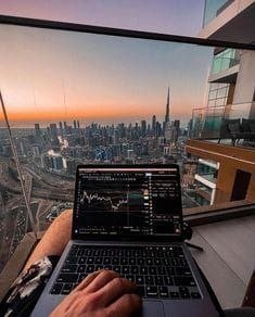 A lifestyle photograph showing hands typing on a laptop displaying financial charts, with a stunning view of the Dubai skyline (including the Burj Khalifa) visible through a large window at sunset.