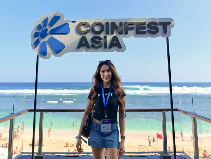 A woman wearing a black 'KIAST' t-shirt, denim skirt, and a lanyard stands on a balcony at a beach, posing beneath a large 'COINFEST ASIA' sign with the ocean in the background.