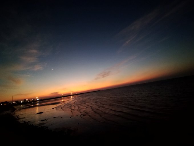 A serene beach at twilight with the sun setting on the horizon, reflecting on the wet sand and water.
