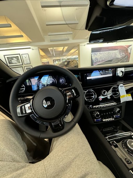 The interior view of a luxury car showing the steering wheel with an 'RR' (Rolls-Royce) logo, dashboard displays, and controls in a showroom setting.
