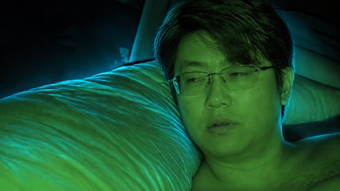 A man with glasses resting his head on a pillow in a dimly lit room, illuminated by a green and blue glow, appearing to be in a scene from a film or TV show.