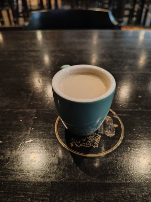 A blue ceramic cup filled with milky coffee sits on a decorative coaster atop a dark wooden table.