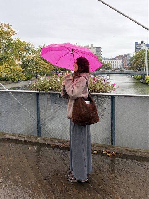 A woman stands on a wet wooden deck holding a bright pink umbrella, wearing a beige jacket, long gray skirt, and carrying a brown bag, with a river, bridge, and cityscape visible in the background on an overcast day.