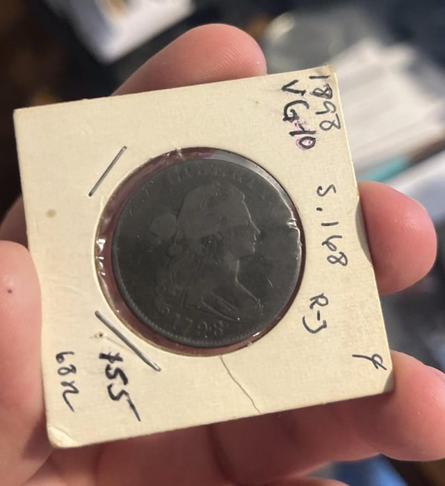 A hand holding an old, dark copper coin (dated 1798) inside a paper flip with handwritten notations including '18728', 'VG10', 'S.168', '27', 'f', and '$55'.