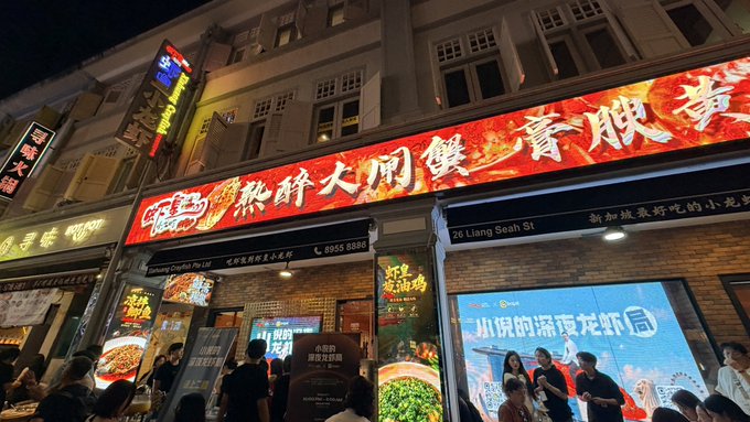 A nighttime street view of a Chinatown shophouse featuring a brightly lit red restaurant sign with Chinese text and a blue advertisement board below it, with crowds of people on the pavement.