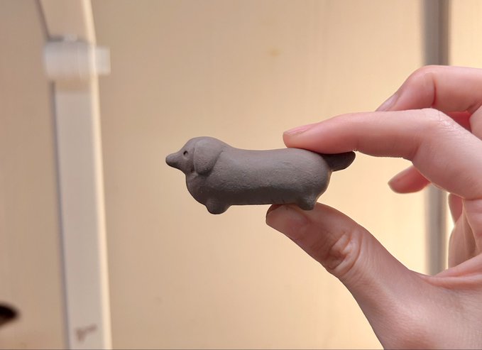 A hand holds a small clay sculpture of a dachshund with notably short legs relative to its long body, humorously illustrating the caption "Long legs short DOG" by highlighting the breed's signature elongated-but-low profile.