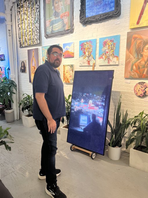 A man with glasses standing in an art gallery next to a large vertical canvas depicting a city nightscape, with various other colorful paintings displayed on the white brick wall behind him.