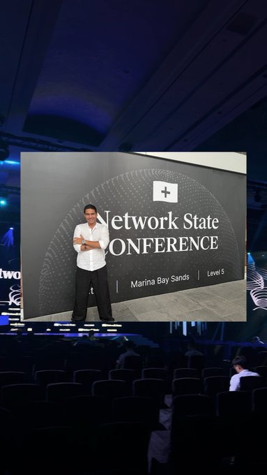 A conference scene showing a man standing on stage in front of a large screen reading 'Network State CONFERENCE, Marina Bay Sands, Level 5,' with an audience seated in a dark auditorium.