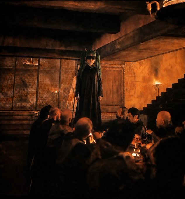 A masked figure in a long black coat stands among a crowd seated at a long table in a dimly lit, cavernous room lit by firelight.