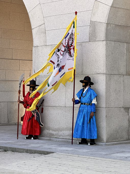 Two Korean guards in traditional red and blue ceremonial robes stand holding a large banner featuring a tiger design and Chinese characters, representing the historic changing of the guard ceremony at Gwanghwamun Gate in Gyeongbokgung Palace. This iconic cultural spectacle in Seoul sets the stage for the author's upcoming return to Korea for the KBW (Korea Blockchain Week) conference.