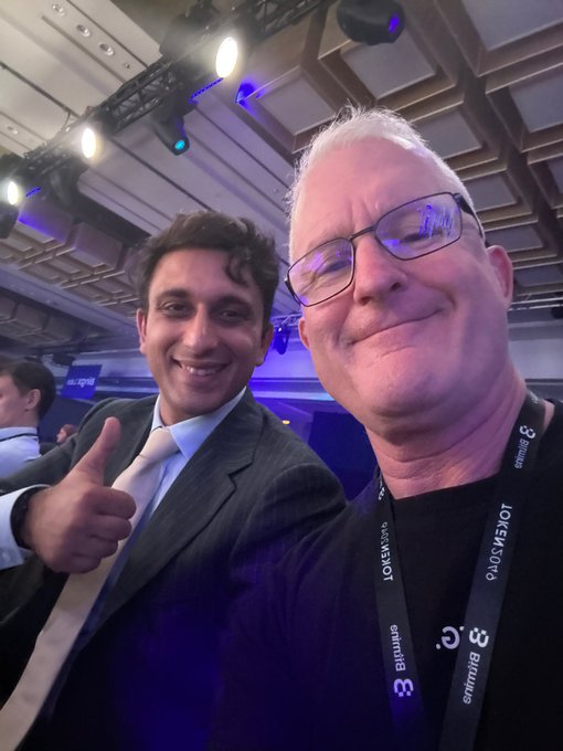 Two men wearing lanyards (one labeled 'LOTUS/SAFE', another 'Bitmain') pose for a selfie at a tech conference with blue lighting.