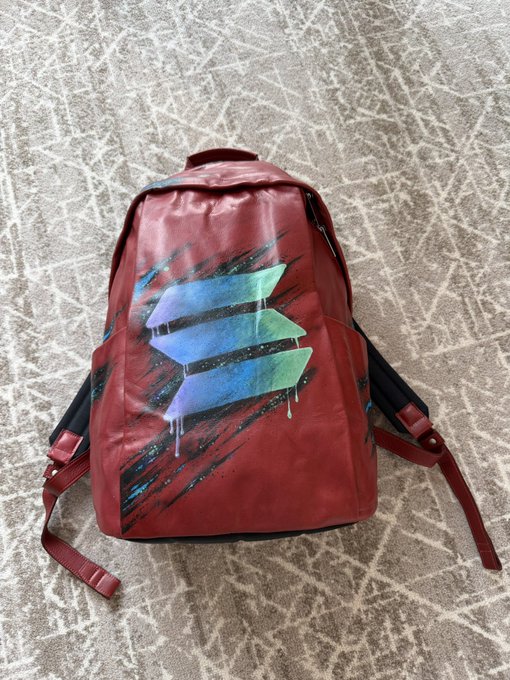 A red backpack with a unique abstract design featuring blue and green geometric shapes with paint drips, photographed lying on a textured concrete surface.