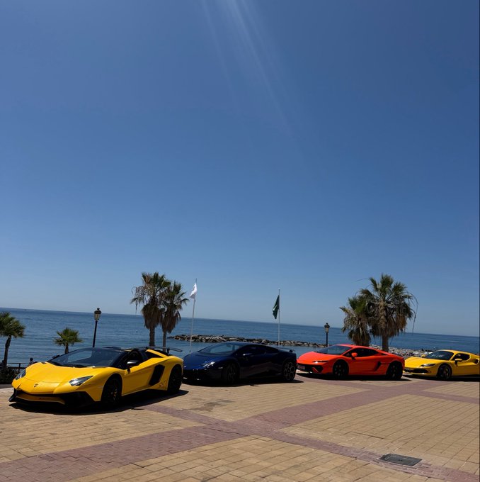 A convoy of sleek Lamborghinis—two yellow, one black, and one orange—are parked along a sunny coastal promenade in Spain, with palm trees and the Mediterranean Sea in the background, fitting the post's proud boast of being a Lamborghini owner who sold at the top.