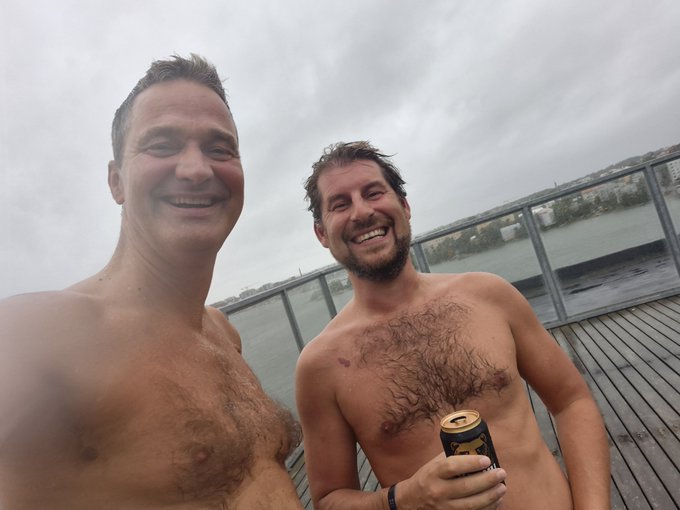 Two shirtless men smile for a selfie on a wooden deck by a body of water, one holding a beverage can.