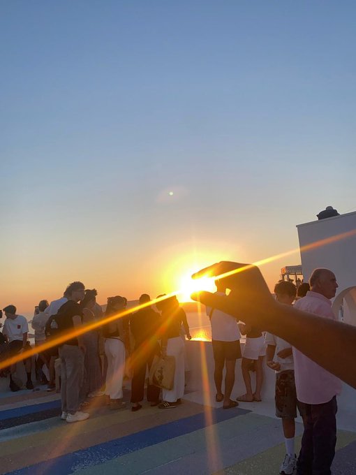 A sunset photo captures a crowd of onlookers gathered as someone playfully holds up their hand to "catch" the descending sun between their fingers, with the bright orb glowing intensely and creating dramatic lens flares across the coastal scene. The hand-sun illusion humorously connects to the post's joke about catching the "next runner" and questioning whether the gesture represents generational wealth or just getting dramatically burned by the sun.
