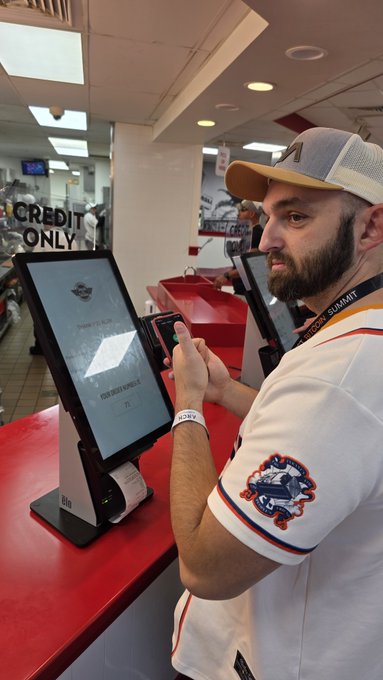 A bearded man wearing a 'Director's Summit' lanyard and Miami-themed shirt swipes a card at a kiosk terminal in a credit-only store.