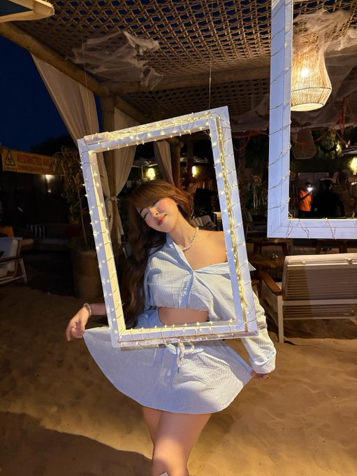 Creative photo of a woman in a white striped outfit posing through a decorative white picture frame wrapped in fairy lights at a beachside venue.