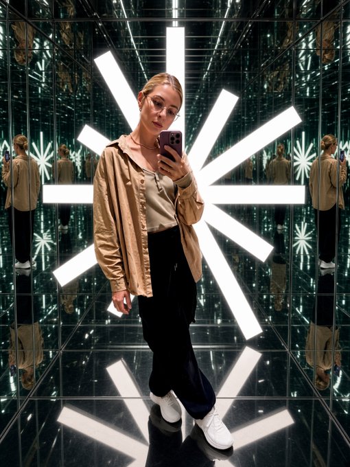 A woman with glasses and a tan jacket takes a mirror selfie in a room with reflective surfaces and bright star-shaped lights in the background.