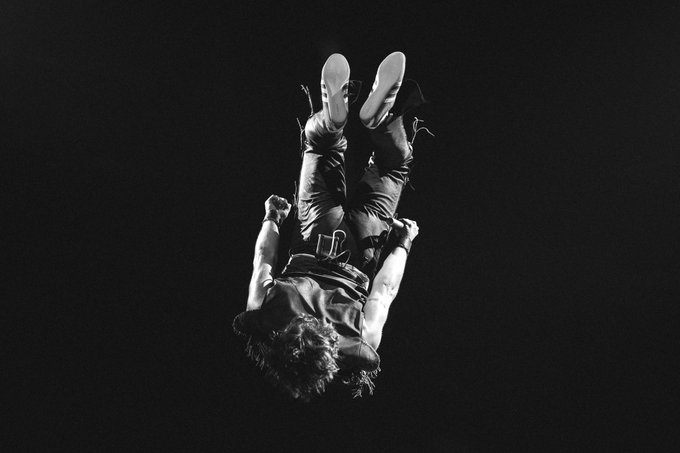 A black-and-white concert photo captures Benson Boone mid-air during a dynamic stage performance, his body inverted with feet pointed skyward against a dark background, perfectly embodying the high-energy live show referenced in the tour announcement.