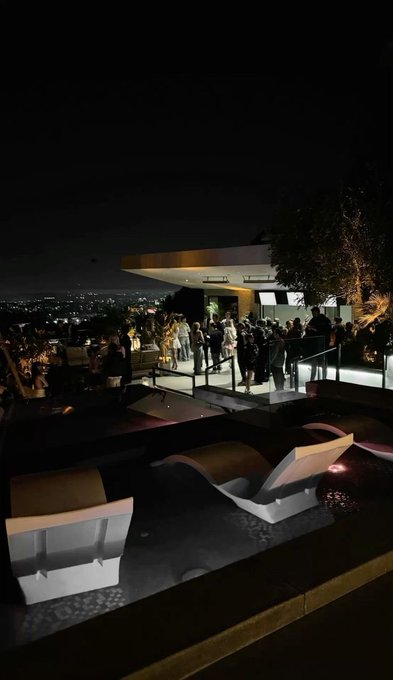 A nighttime party scene on a hilltop with a crowd of people, illuminated by city lights in the distance, and modern white lounge chairs beside a pool.