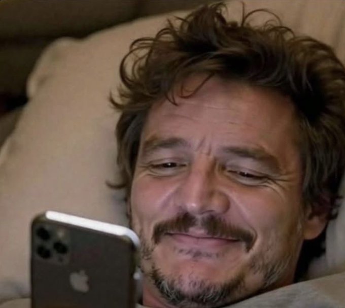 A close-up photo of a smiling man with curly brown hair and a mustache lying down while looking at an iPhone, with the Apple logo visible on the back of the device.