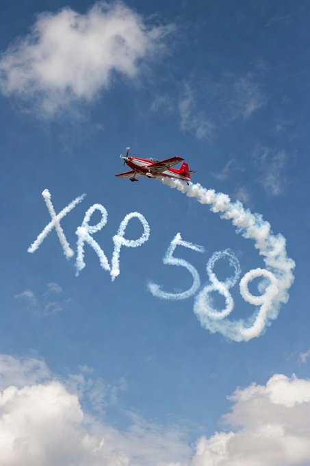 The image features a red airplane flying across a blue sky, leaving behind a trail of smoke as it moves through the air. Below the plane, there are clouds floating around. On the right side of the image, the words "XRP" are written in white letters that appear to be spray painted or drawn by someone, possibly at an air show or similar event where airplanes fly low for spectators to see up close. The combination of the airplane and smoke creates a sense of movement and excitement, reflecting the dynamic nature of this form of aerial acrobatics.