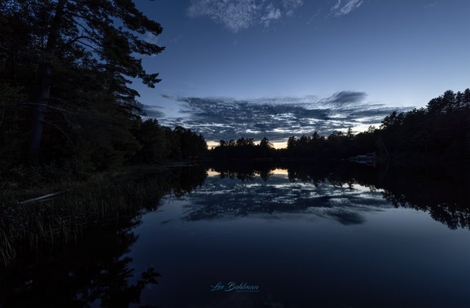 A serene twilight photograph captures a calm lake reflecting the blue sky and scattered clouds, framed by dark silhouettes of pine trees and foliage, with a photographer's watermark 'Joe Bohman PHOTOGRAPHY' visible at the bottom.