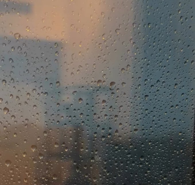 A close-up view through a rain-covered window, showing water droplets on the glass with a blurred city skyline and clouds visible in the background.