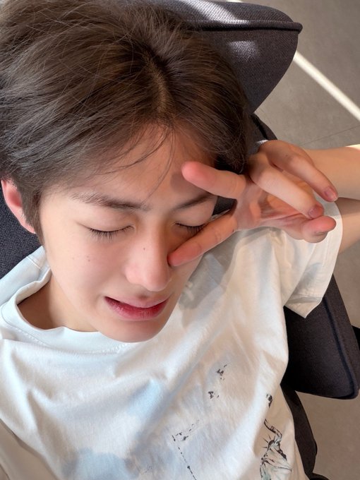 Close-up selfie of a young man with dark hair wearing a white t-shirt, eyes closed with his hand making a peace sign near his face