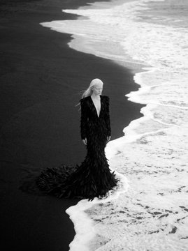 A black-and-white image of a blonde woman in a long, textured black gown standing at the edge of the ocean where waves meet the shore.