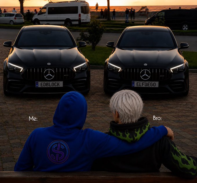 The image shows two men sitting next to each other, both wearing hoodies. They appear to be enjoying a moment together as they look at their cars parked nearby. The background of the photo features a beautiful sunset over a beach or park setting, creating a serene atmosphere for the scene. There are also several people in the distance, possibly enjoying the view and the company of others.