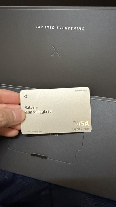A hand holds a sleek, silver Visa debit card personalized with the name "Satoshi" and username "@satoshi_gfa18," presented in front of a black box inscribed with "TAP INTO EVERYTHING" and the "X" logo, visually matching the excitement of receiving the X Money card.