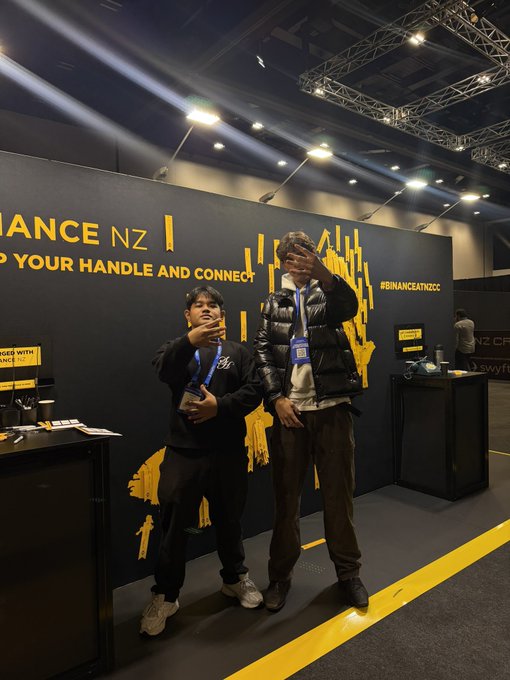The image features two people standing next to each other with their hands behind their backs, possibly posing for a picture or participating in an event related to BINANCE NEW ZEALAND.