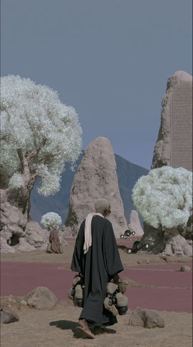 A digitally altered image depicts a person in dark robes carrying jugs, walking through a surreal landscape featuring large rock formations, pale trees, and a reddish ground plane under a clear blue sky.