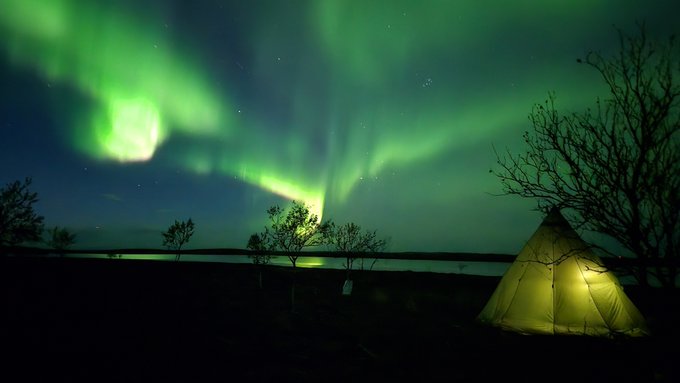 A glowing tent sits in a dark landscape beneath a vivid green aurora borealis lighting up the night sky over a body of water.